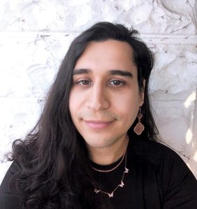 A woman with long brown hair wears a black top and stands in front of a white stone wall.