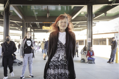 A woman with red hair, wearing a long cardigan and black/grey/white dress stands on a bus platform.
