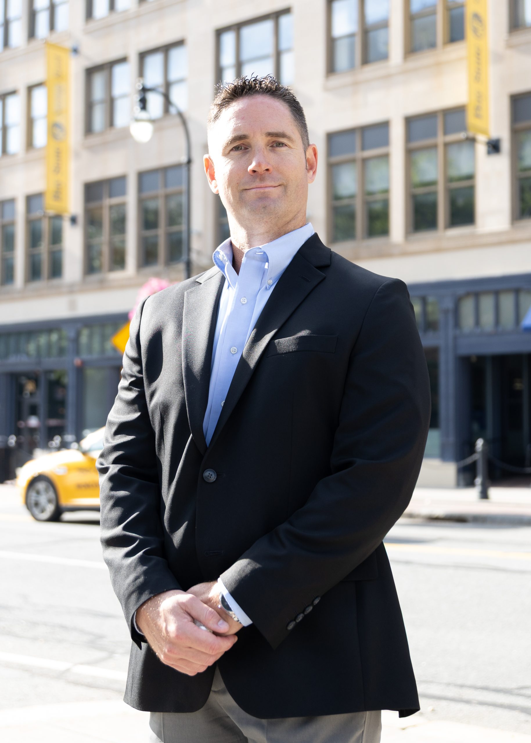 A man wearing a suit jacket stands holding his hands together while standing outside in front of a street.
