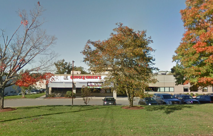 A one-story commercial building sits behind some trees, a lawn, and a parking lot.