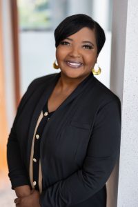 A woman with short black hair wears a long black cardigan and gold earrings.