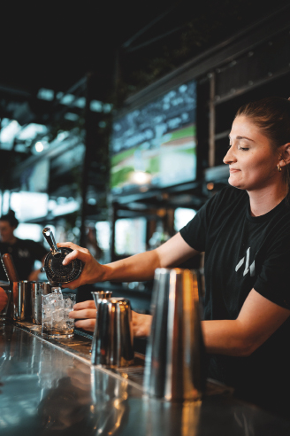 A woman pours a drink behind a dark bar with a tv behind her playing a baseball game.