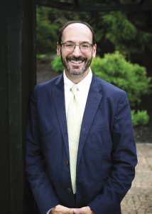 A headshot of a man with brown hair and glasses wearing a blue suit jacket, a white button down, and a light green tie