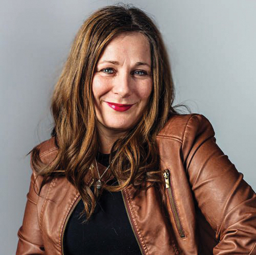 A woman with long brown hair wears red lipstick and a a light brown leather jacket.