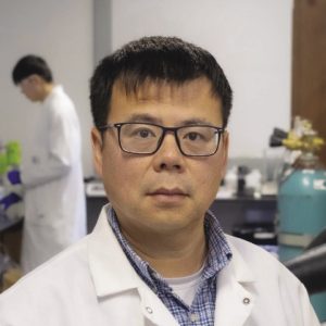 A man wears black glasses and a white lab coat.
