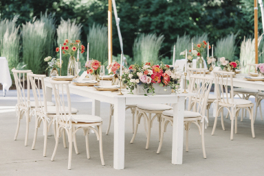 White tables outside decorated with pink flowers in front of green shrubbery 