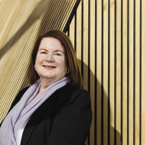 A woman with shoulder-length red hair wears a black jacket blazer and a light purple scarf.
