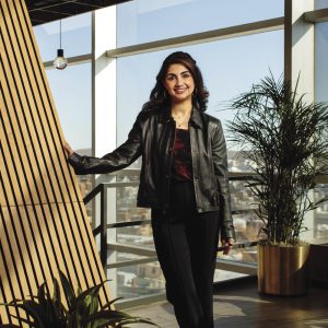 A woman wears a black leather jacket, a red and black top, and black pants in front of a wall of windows.