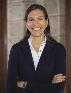 A woman with long brown hair stands with her arms crossed in front of her wearing a white top and blue cardigan