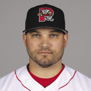 A man wears a baseball cap and a white and red baseball uniform.