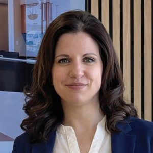 A woman with long brown, waved hair wears a dark blue blazer and a white top.