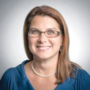 A woman with collar bone-length light brown hair wears a blue top and pear necklace.