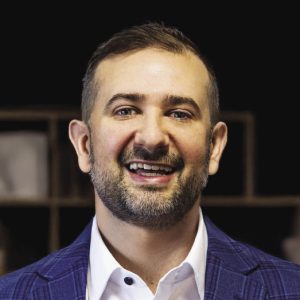 A headshot of a man with short brown hair wearing a dark blue plaid blazer and a white button down