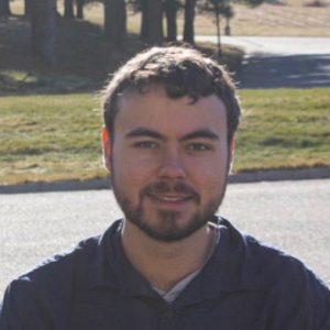 A man with short brown hair sits outside in front of a green area with trees.