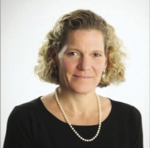 A woman with chin-length blonde wavy hair wears a black top and pearl necklace.