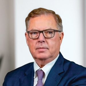 A headshot of a man wearing a dark blue suit jacket, white button down, and purple tie