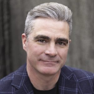 A man with grey hair wears a blue plaid blazer.