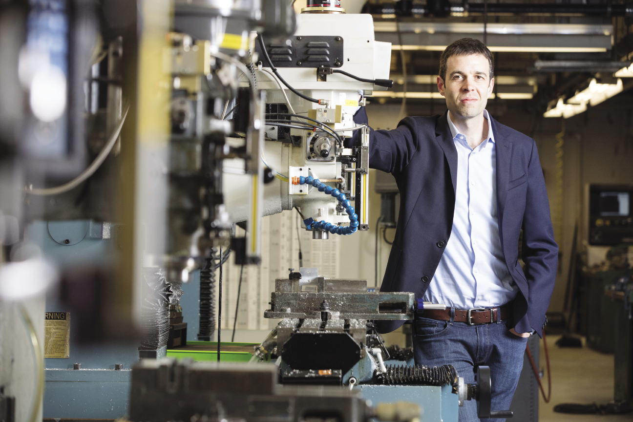 A man wearing a light blue button down, a dark blue suit jacket, and jeans leans against a row of machinery.