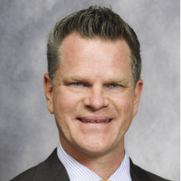 A man smiles for a headshot while wearing a light blue button down shirt and a black blazer.