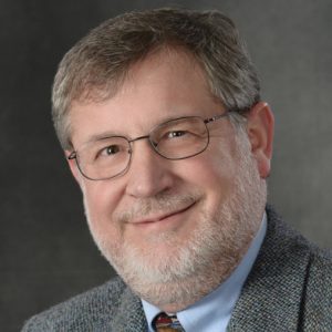 A man with grey hair and beard wears glasses and a grey suit jacket.
