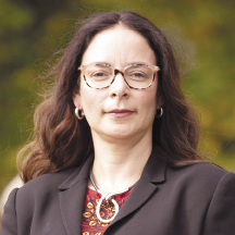 A woman with long, wavy brown hair wears a black blazer and red top.