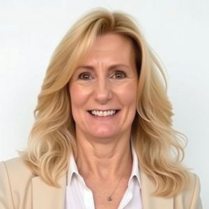 A woman with blonde, wavy hair wears a white button down and a beige suit jacket.