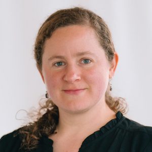 A woman with curly blonde hair tied back wears a black top.