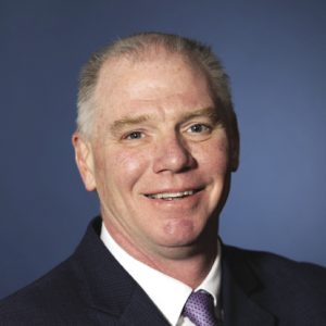 A man with white hair wears a black suit jacket, white button down, and purple tie.