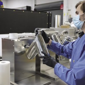 A man wears blue scrubs and a mask while holding a manufacturing tool.