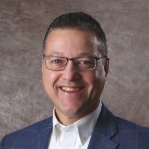 A man wears glasses, a blue suit jacket, and white button down.