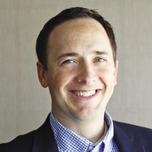 A man wears a dark blue suit jacket and white and blue checkered button down.