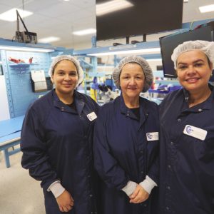 Three women stand together wearing hairnets and dark blue lab coats.
