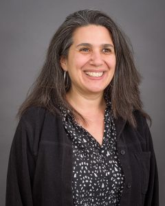 A woman with long, straight grey hair wears a black and white top.