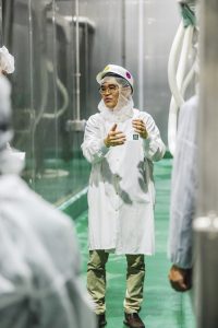 A man wears a white lab coat and safety equipment while standing inside a manufacturing facility.