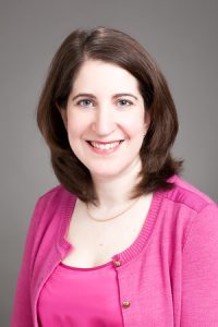 A woman with shoulder-length brown hair wears a pink top and cardigan.
