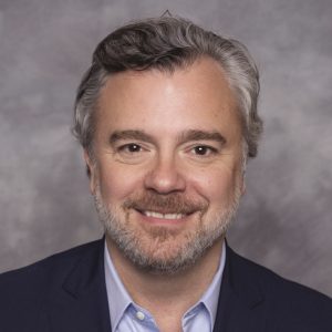 A man with grey and white hair wears a dark blue suit jacket and light blue button down.