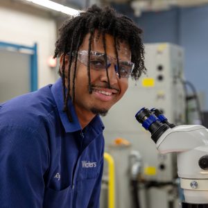 A man with curly dark hair wears a blue polo shirt while leaning towards a microscope.